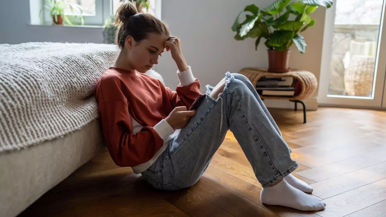 O adolescentă îmbrăcată cu un hanorac roșu stă sprijinită de o canapea în timp ce folosește un smartphone.