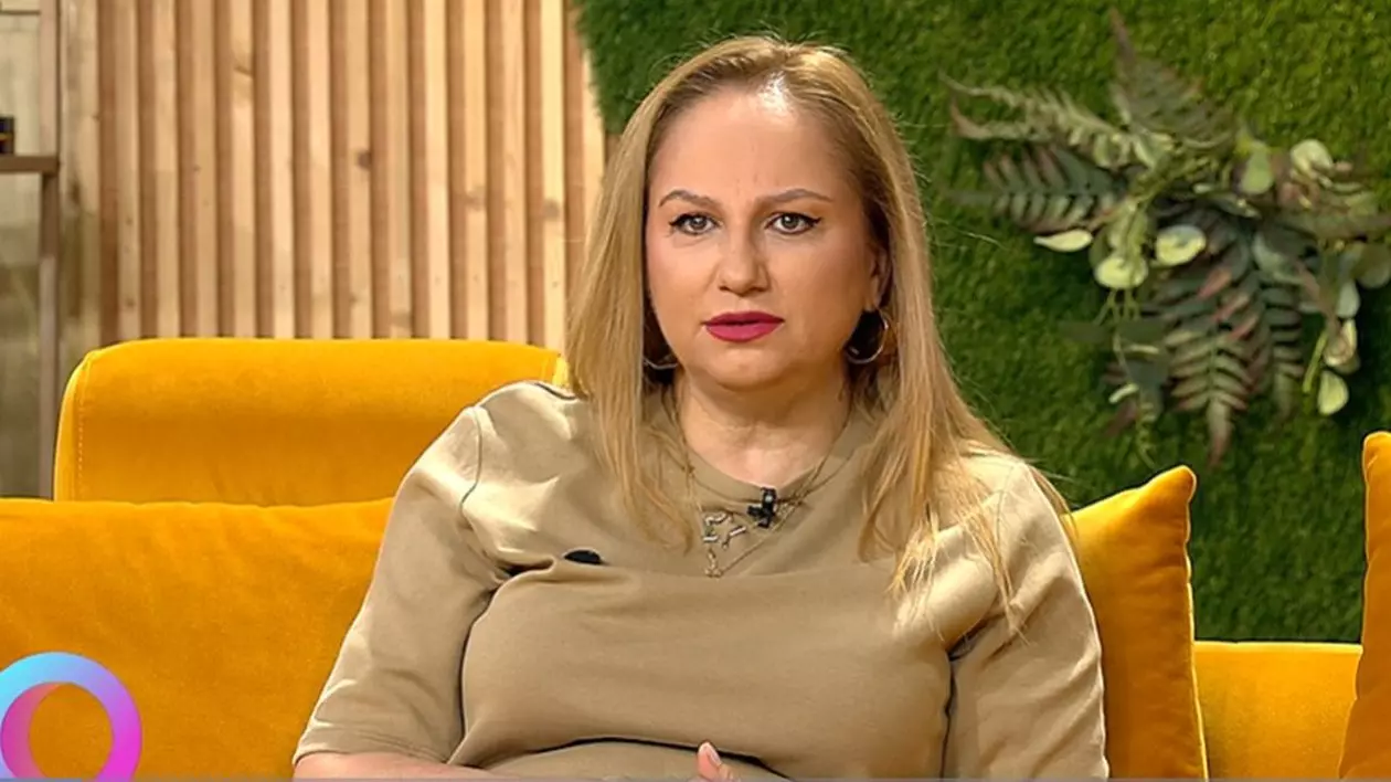 Astrologul Cristina Demetrescu, îmbrăcată cu o bluză maro, stă pe o canapea portocalie în studioul unei emisiuni TV