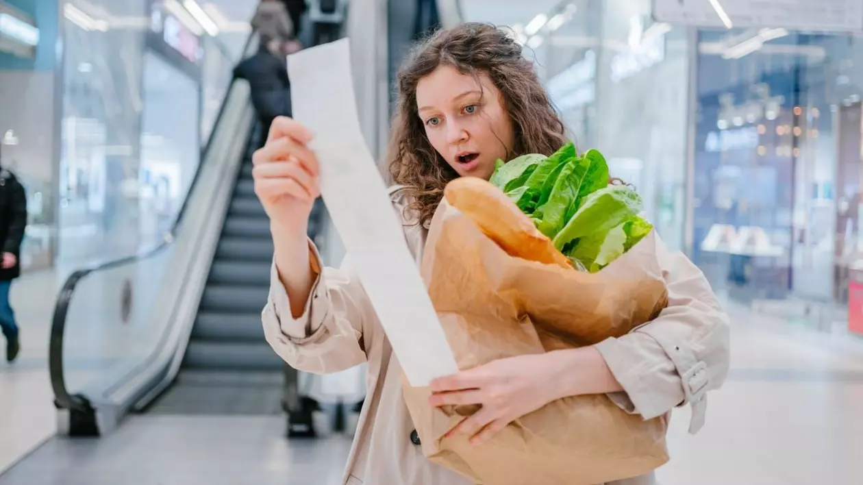 O femeie se uită foarte mirată la un bon de cumpărături într-un supermarket dintr-un mall și are în brațe un pachet cu pâine și salată, iar în fundal se văd scări rulante