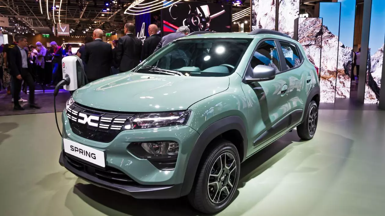 Model Dacia Spring electric, de culoare verde deschis, expus într-un showroom auto. În fundal sunt mai multe persoane.