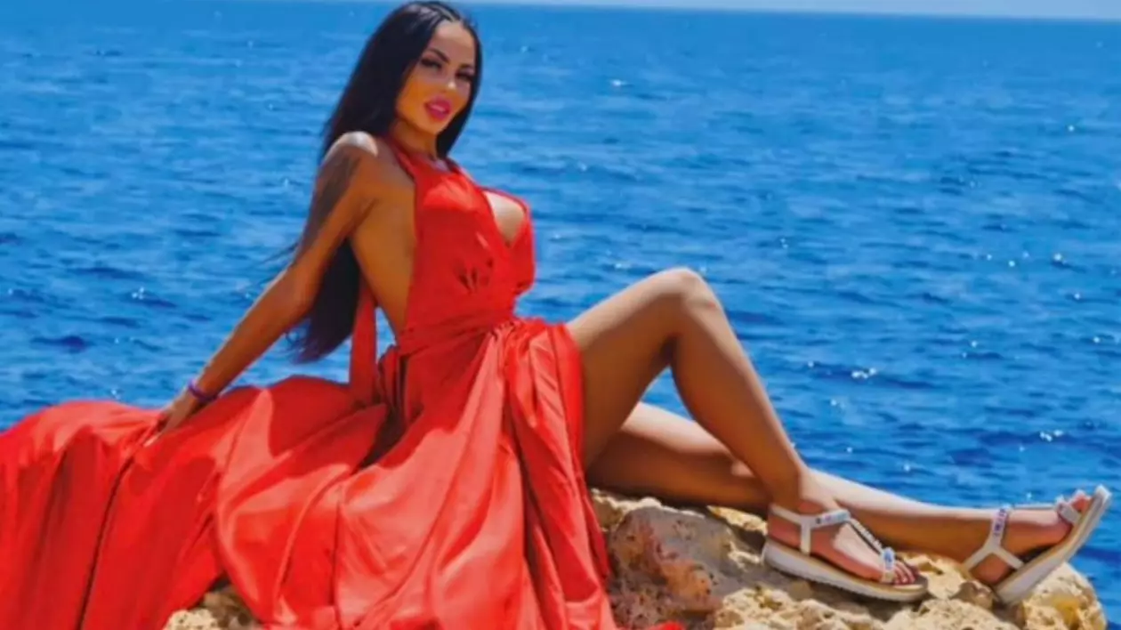 Denisa Maria Adas, într-o rochie roșie cu trenă, în timp ce stă pe o piatră, cu marea pe fundal