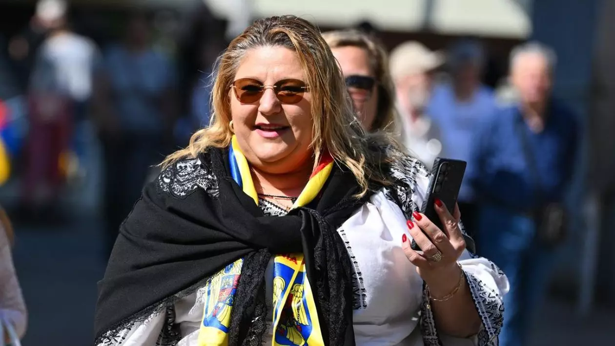 Diana Sosoacă intr-o ipostază pe stradă, rade, cu telefonul în mana stanga, care este ridicata, poartă o bluză alba cu model traditional romanesc si o eșarfă neagră legata la gat, oameni in spatele ei