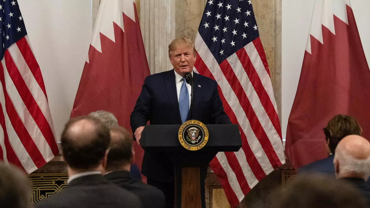 Donald Trump, la o conferință de presă, îmbrăcat cu sacou negru, cămașă albă și cravată albastră, la un pupitru. În spatele lui sunt steagurile Americii și al Quatarului.