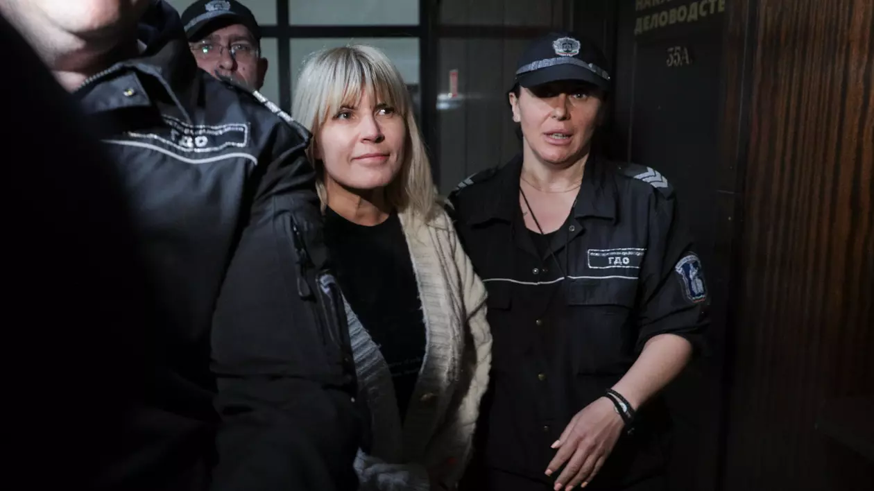 Elena Udrea, îmbrăcată cu o bluză neagră și o jachetă crem, cu părul desfăcut, fiind însoțită de o polițistă și doi polițiști îmbrăcați în uniforme de vară cu cămăși bleumarin cu mânecă scurtă