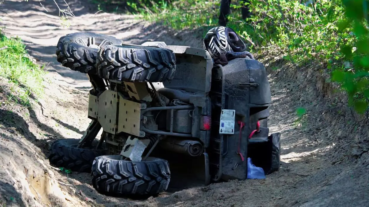 Un ATV negru răsturnat pe partea dreaptă pe un drum din pădure, cu iarbă verde în jur