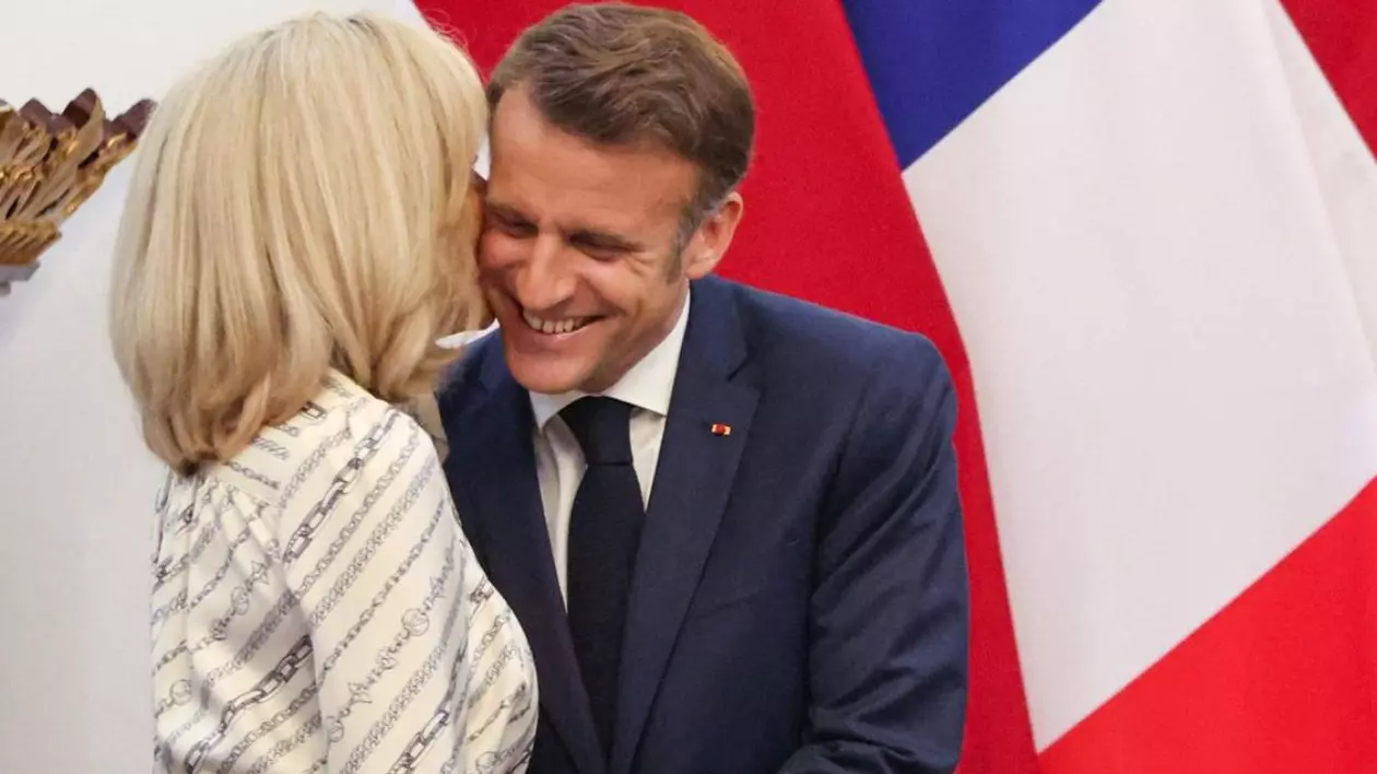 Emmanuel Macron, in costum inchis la culoare si camasa alba, zâmbește în timp ce soția sa Brigitte, intr-o rochie crem cu model, sta cu spatele si ii spoteste ceva la ureche presedintelui
