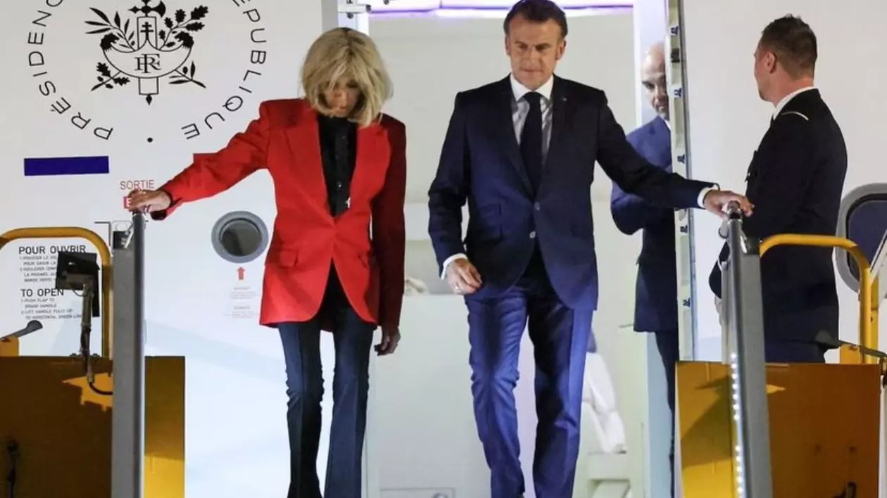 Emmanuel Macron, in costum inchis la culoare. se tine cu mana stangă de bara scării de la avion, și Brigitte Macron, în sacou rosu su pantalon negru, se tine cu mana dreapta de bara de la scara avionului, coboara din aeronava