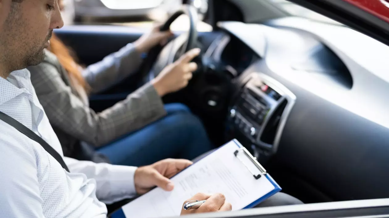 În imagine este ilustrat momentul examenului pentru permisul auto. O tânără se află la volan, iar examinatorul în partea dreaptă.