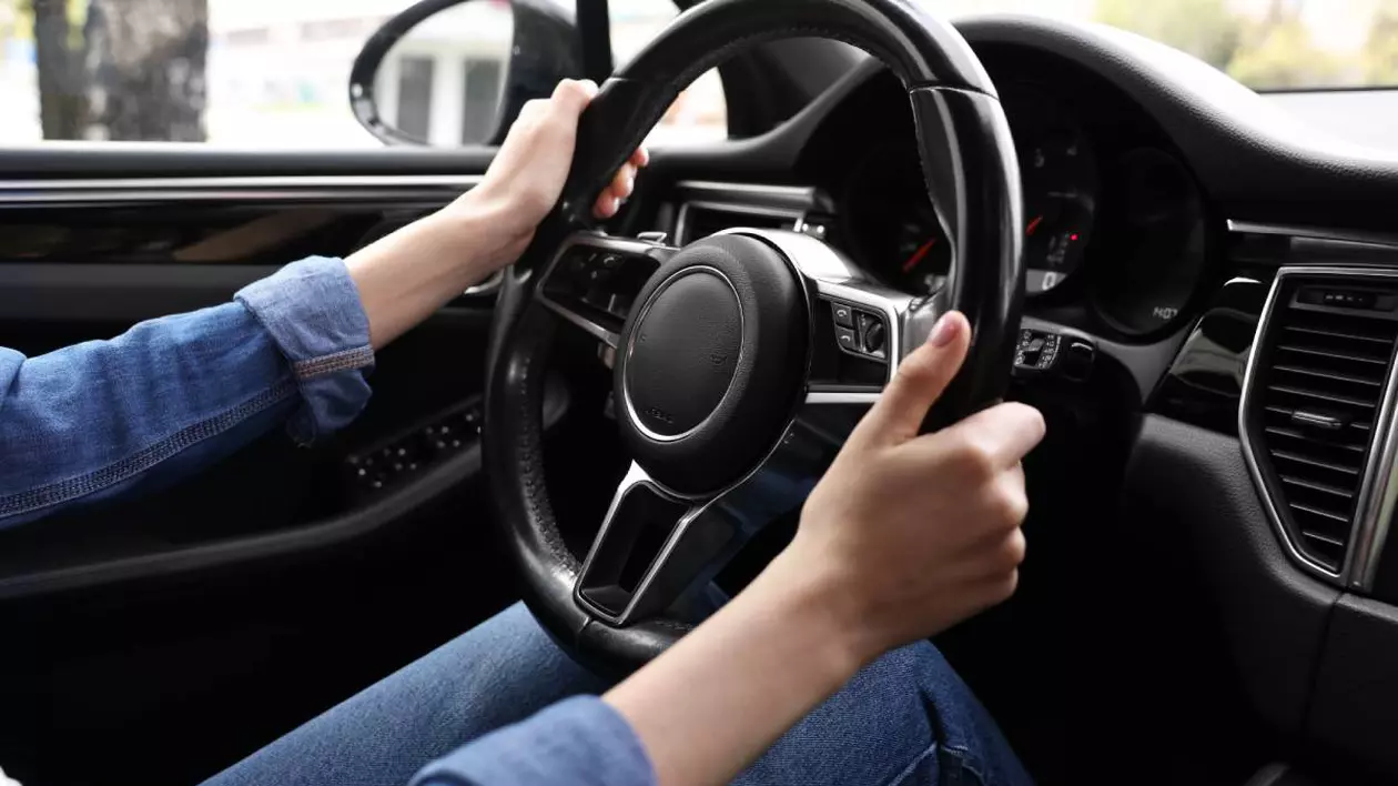 Șofer aflat la volanul unei mașini, care ține cu ambele mâini pe volan. Persoana poartă o cămașă de blugi și blugi albaștri. Bordul mașinii este unul modern