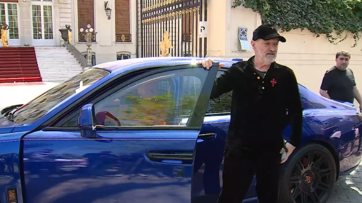 Gigi Becali, îmbrăcat într-un trening de culoare neagră, inscripționat cu o cruce roșie, cu o șapcă neagră pe cap, în timp ce se dă jos dintr-un Rolls Royce de culoare albastră