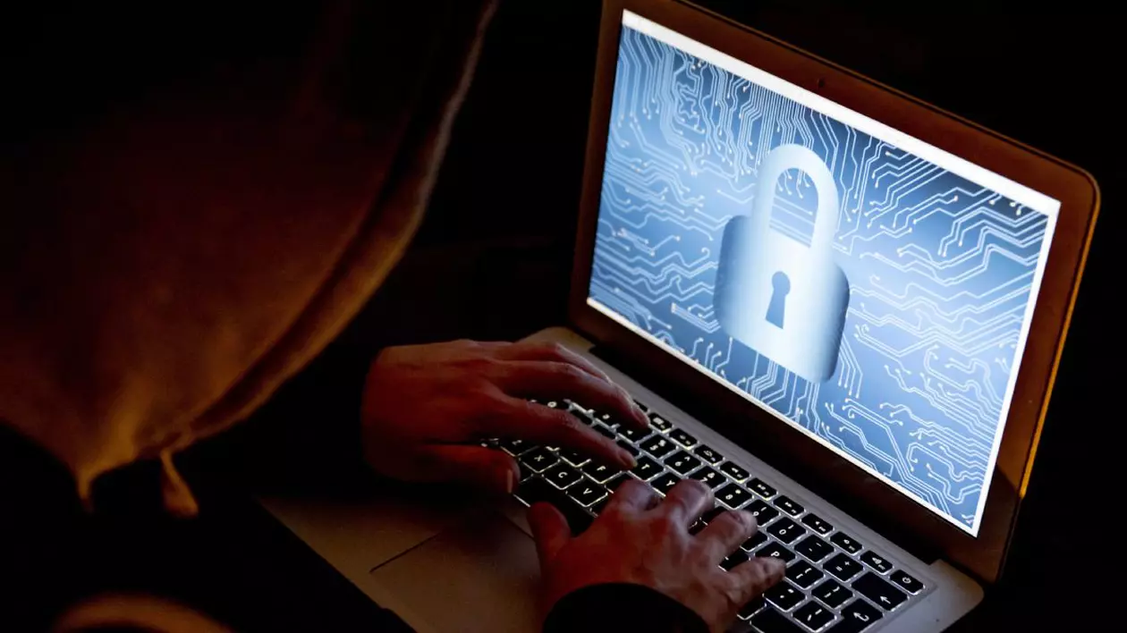 Un bărbat cu glugă care seamănă cu un hacker lucrează la un laptop care are afișat pe ecran un lacăt pe funal albastru deschis.