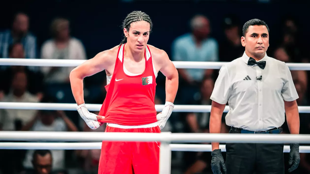 În imagine este o persoană îmbrăcată în haine roșii, într-un ring de box. Lângă aceasta se află un bărbat în cămașă albă.