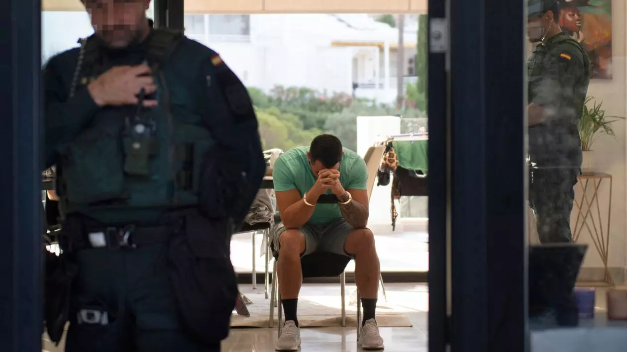 În prim plan un polițist spaniol îmbrăcat în uniformă, iar în plan secund un infractor cu cătușe la mâini, care stă pe un scaun, îmbrăcat în tricou și pantaloni scurți