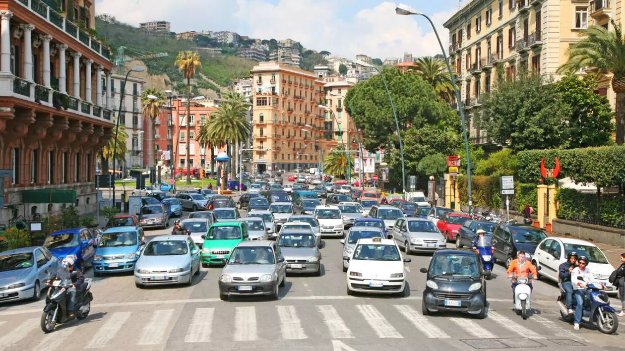 Trafic urban intens într-un oraș din Italia, într-o zonă centrală cu clădiri istorice, palmieri și bulevarde aglomerate. Zeci de mașini – în special modele mai vechi, inclusiv diesel – ocupă toate benzile unei intersecții mari