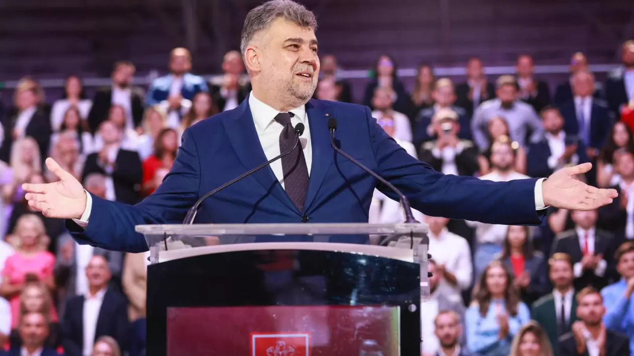 Marcel Ciolacu, îmbrăcat la costum, cu o gravată vișinie, în timp ce participă la un congres al PSD. Liderul socia - democrat stă cu mâinile întinse și zâmbește