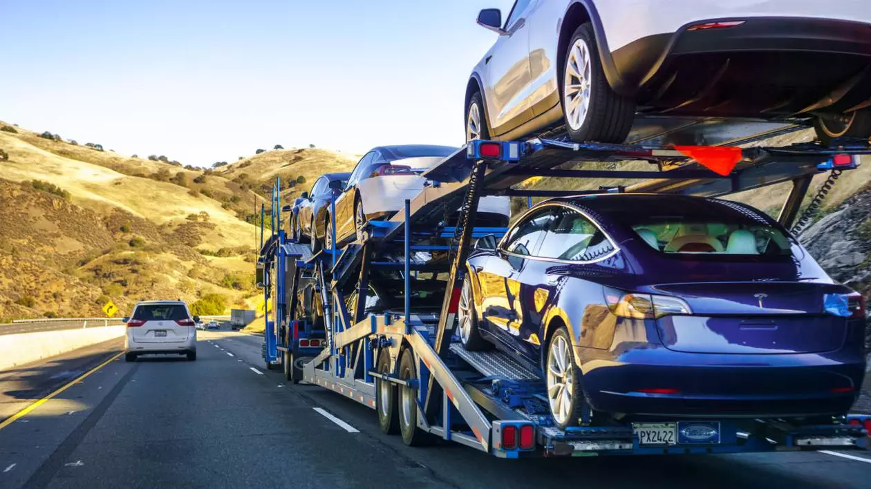 Transport auto pe o autostradă, în care mai multe mașini Tesla sunt încărcate pe o platformă specială cu două niveluri. Vehiculele sunt noi, fără plăcuțe de înmatriculare.