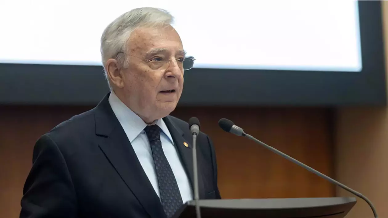 În imagine este ilustrat Mugur Isărescu. Acesta este îmbrăcat în costum negru, cămașă albă și se află în fața a două microfoane.