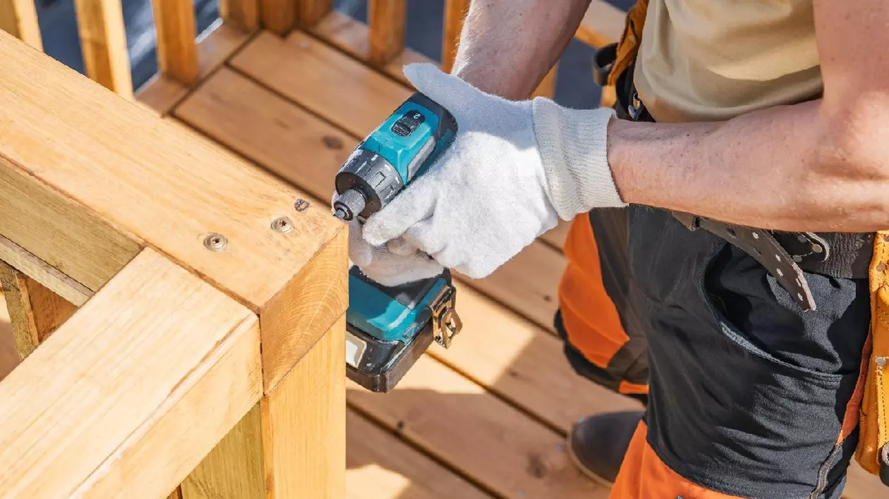 Un muncitor se concentrează pe fixarea unei balustrade din lemn cu un burghiu fără fir într-o zi însorită și luminoasă. Purtând mănuși și pantaloni de lucru portocalii, se asigură cu atenție că balustrada este stabilă pe un șantier de construcții.