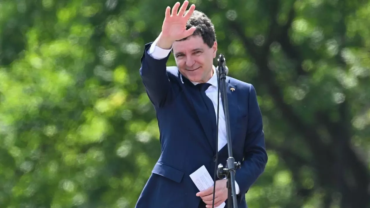 Nicușpr Dan, într-un costum inchis la culoare si o camasa alba, cu mana dreapta ridicata in semne de salut si cu niste foi rulate in mana stanga, într-o zi insorita, cu un copac verde in fundal