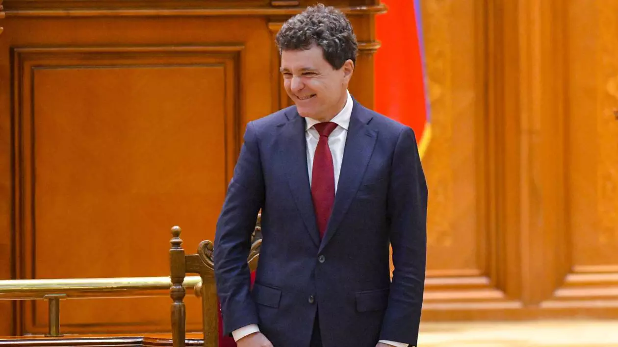 Nicușor Dan în Parlamentul României, în Camera Deputaților, în timpu ce zâmbește și stă cu mâinile pe lângă corp. Are un costum albastru și o cravată roșie.