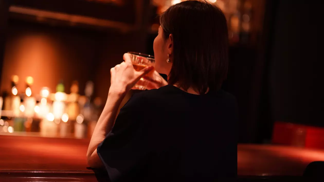 În imagine este ilustrată o femeie care ține un pahar cu alcool în mână, în timp ce stă la un bar