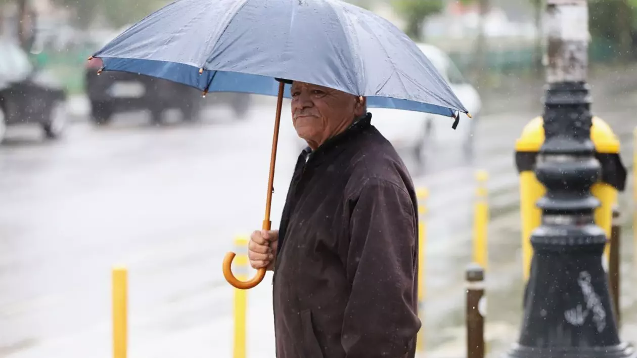Un bărbat în vârstă, cu geacă maro și o umbrelă albastră pe care o ține deschisă, stă la semafor într-o zi ploioasă, marți, 27 mai 2025, București