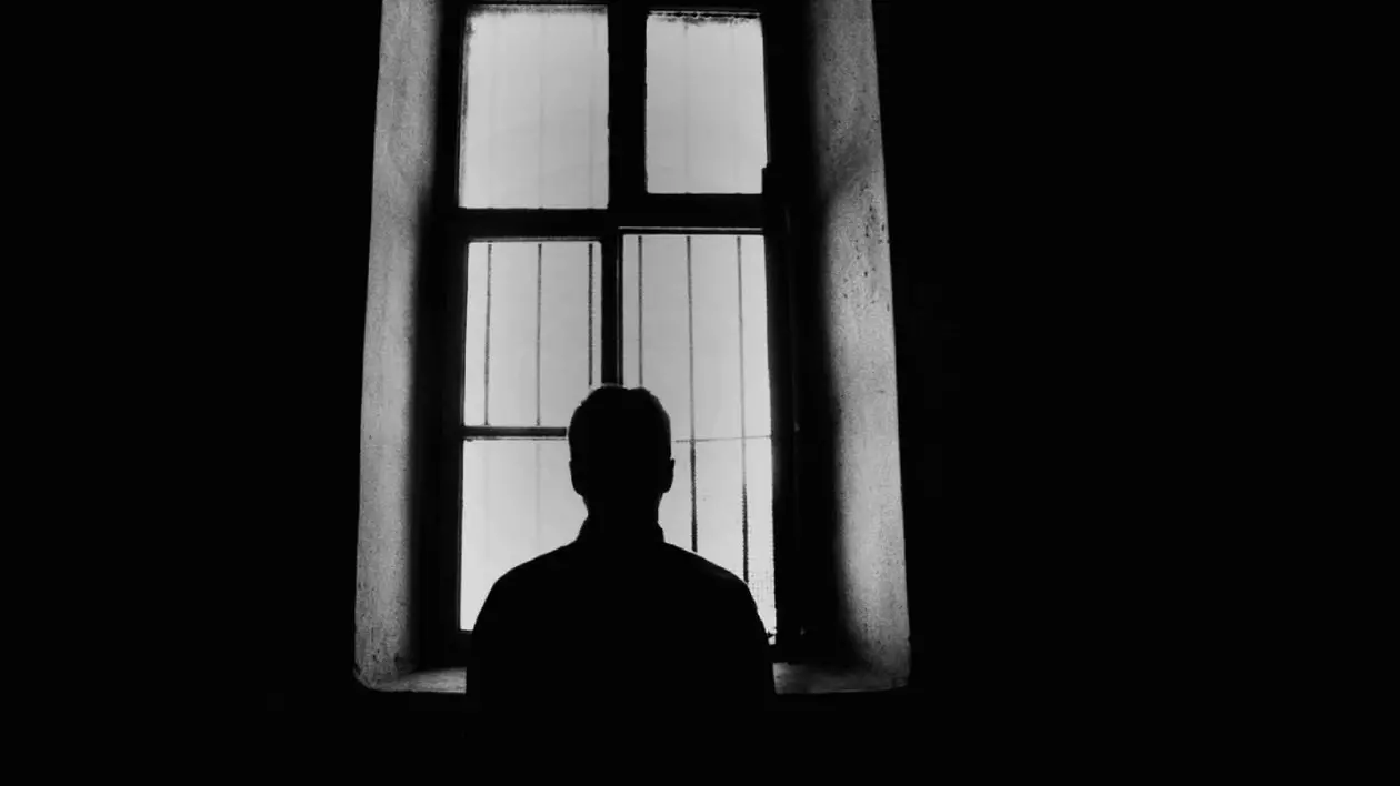 Silueta unui barbat care se uita pe geam, într-o cameră întunecată.