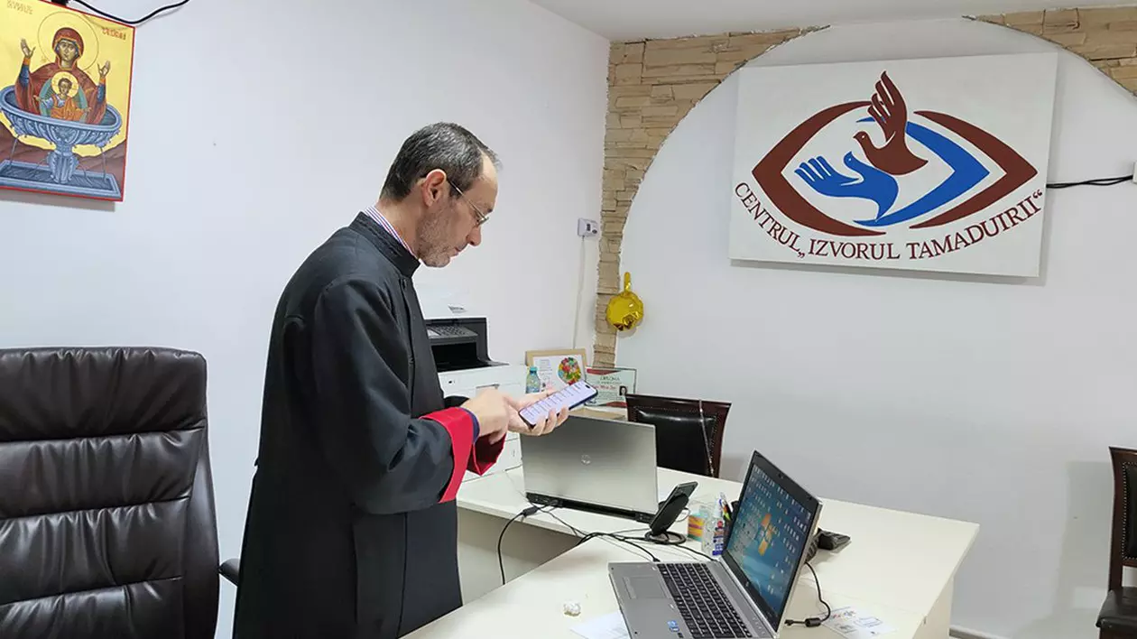 Părintele Liviu Burlacu, centrul Izvorul Tamaduirii, intr-un birou, in fata a doua laptopuri