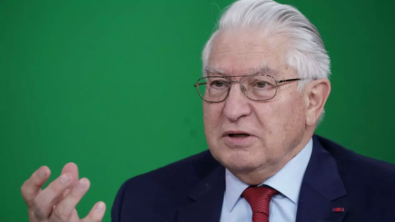 Profesorul dr. Vlad Ciurea, îmbrăcat în costum de culoare albastru închis, cravată roșie și cămașă albă, pe fundal verde, cu o mână ridicată