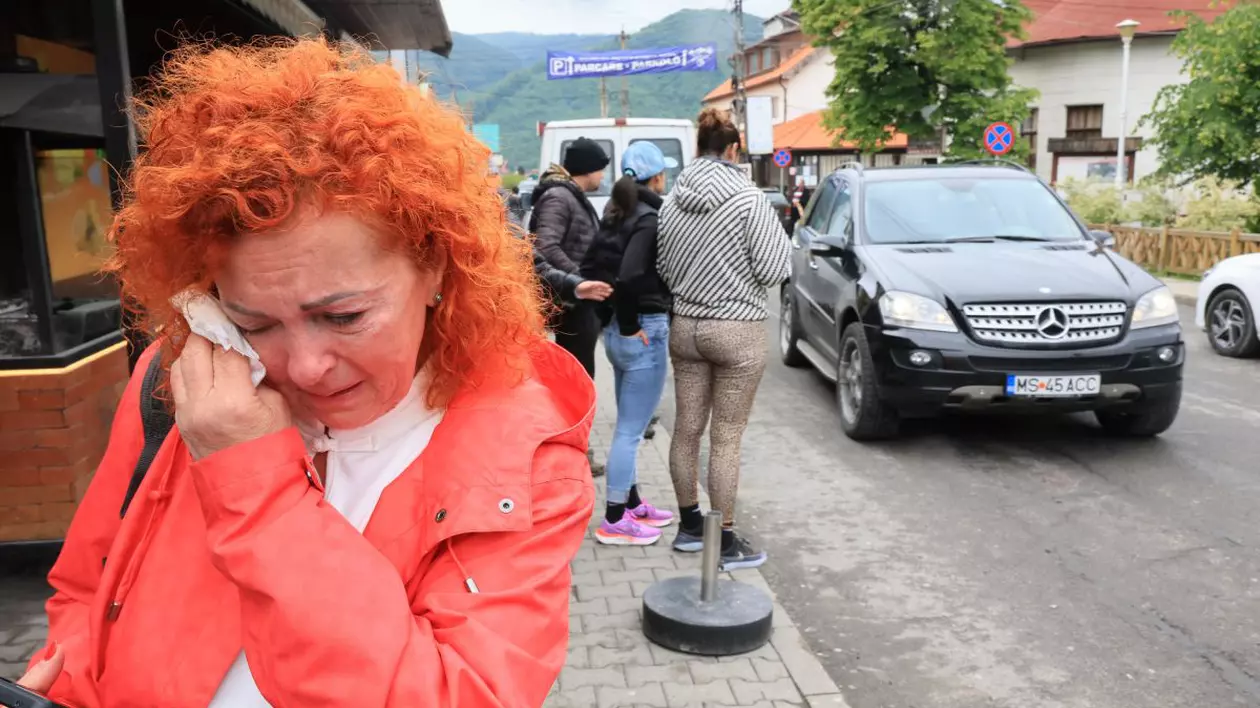 o femeie cu parul roscat si geaga rosie plange si se sterge la ochi cu mana stanga, in spatele ei alte persoane si o masina neagra