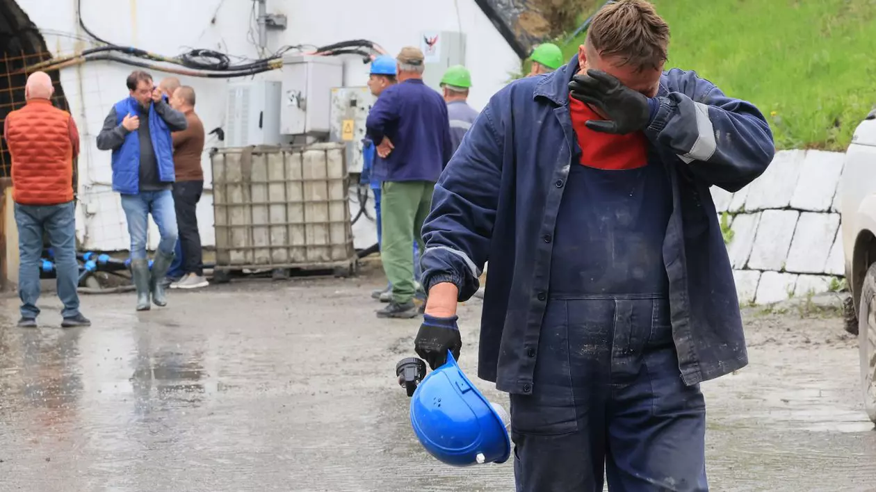 Oameni in uniforma de miner, cu casca si cizme, in fata unei mine, in prim plan un barbat cu mana stanga ridicata la fata si o casca albastra in mana dreapta
