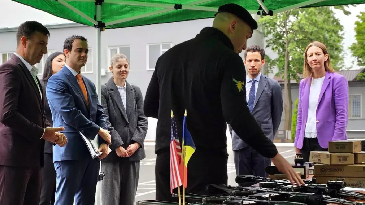 Un polițist se uită la mai multe arme întinse pe o masă, în spate sunt mai mulți oameni, îmbrăcați în costume de diverse culori