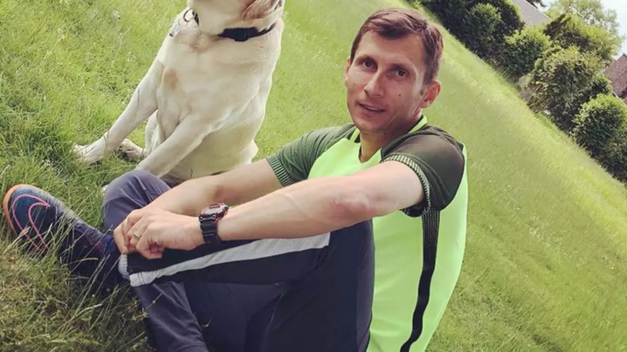 Un barbat stă pe iarbă, într-un parc. Este îmbrăcat cu pantaloni negri și un tricou galben fosforescent de sport. Lângă el stă un câine labrador.