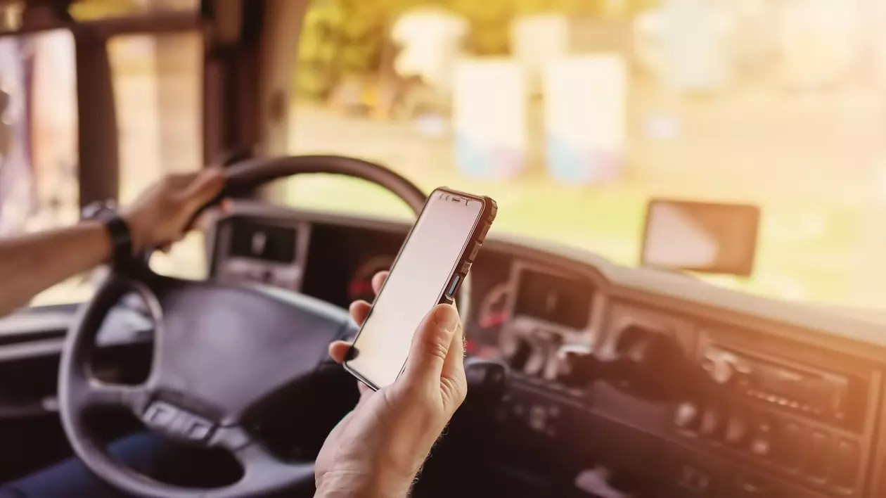 Poză cu un șofer de TIR care ține o mână pe volan, în timp ce în cealaltă are un telefon mobil