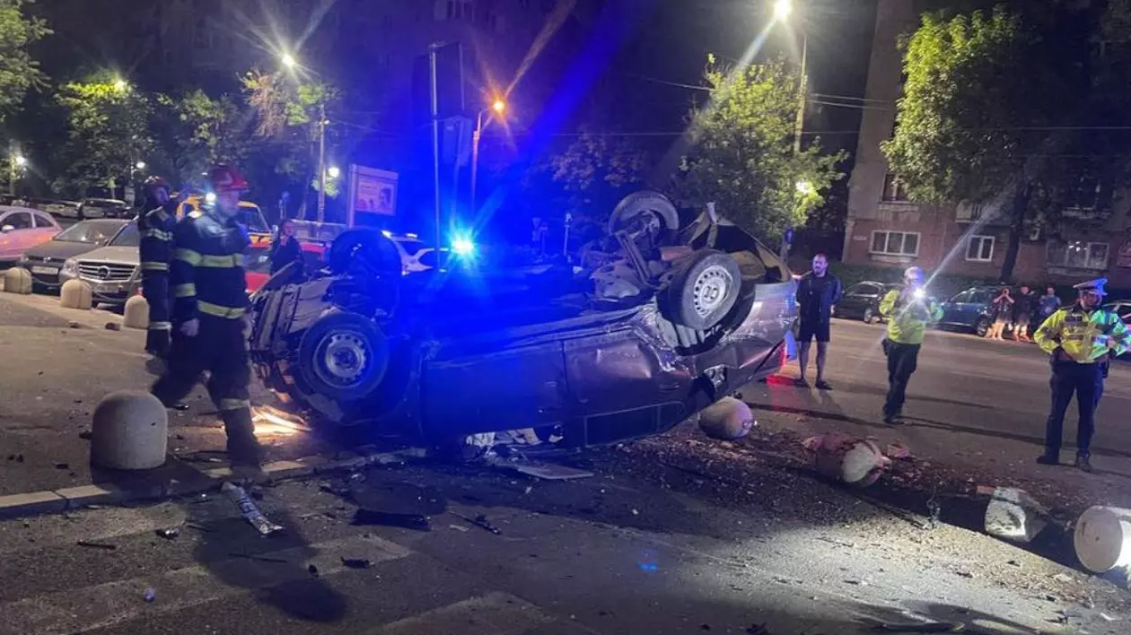 In imagine o masina rasturnată, distrusă, politie si pompieri in jurul ei, in fundal lumini de girofar, noaptea