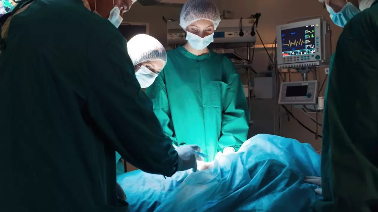 Medici chirurgi, imbracati in costume verzi si cu bonete pe cap, in sala de operatie, aplecati asupra unui pacient