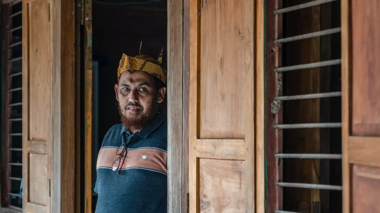 Un bărbat stă în ușa unei case din lemn. Decorul rustic sugerează un cadru cultural local sau rural.