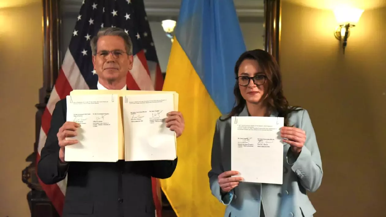 Un bărvbat și o femeie țin în mână și arată câte un document. Bărbatul este îmbrăcat în costum negru și are ochelari, iar femeia într-un costum bleu deschis. În spate sunt steagurile Ucrainei și cel al Statelor Unite