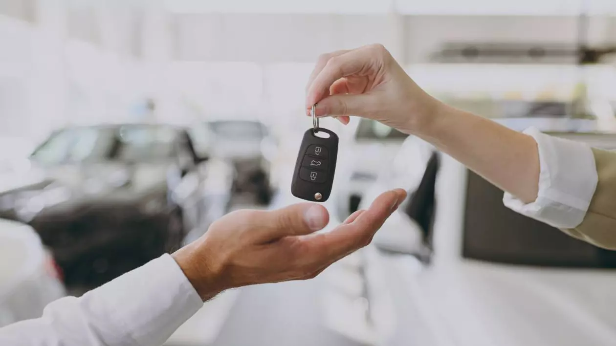 O persoană înmânează cheia unei mașini către un nou proprietar. Imaginea este realizată într-un showroom auto, cu vehicule în fundal ușor blurate.