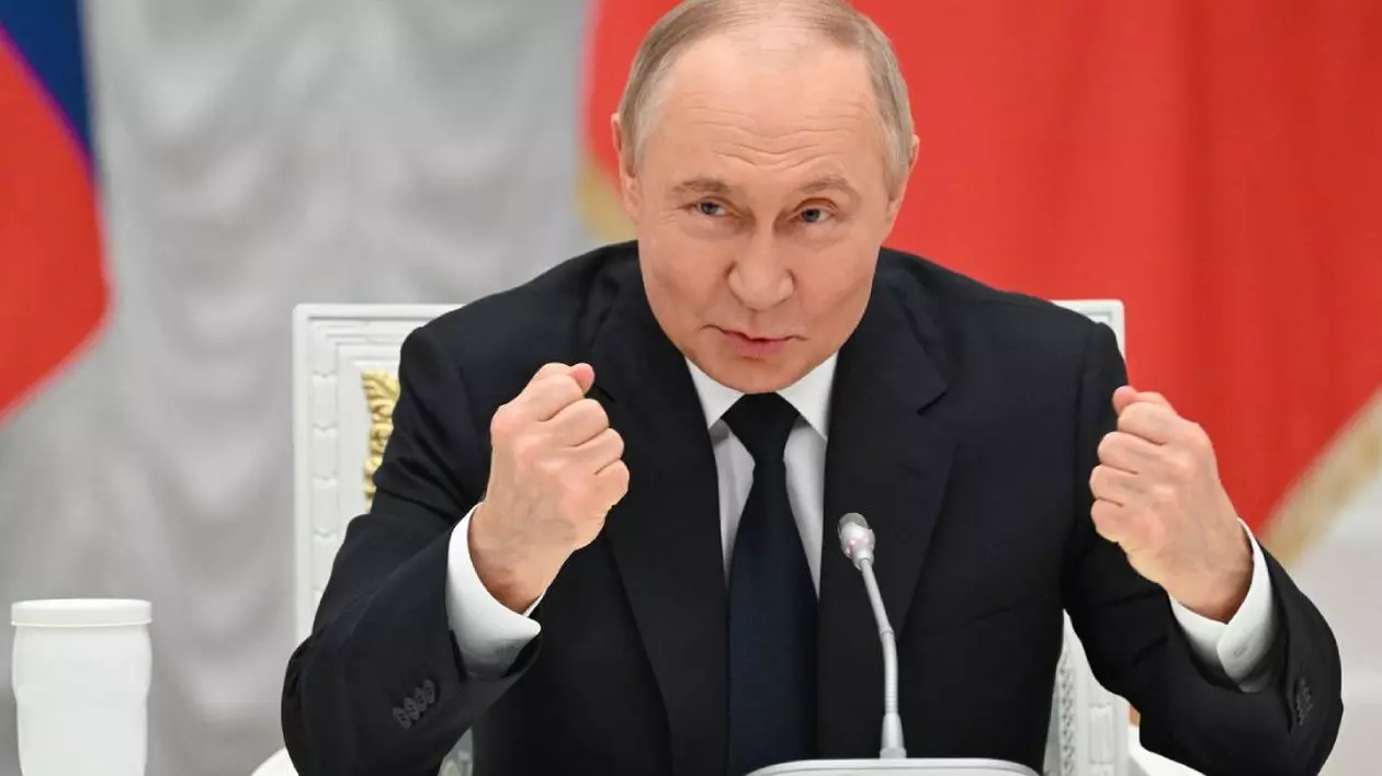 În imagine liderul rus Vladimir Putin, cu mâinile ridicate și pumnii strânși, stând la masă, zâmbind, pe un fundal roșu