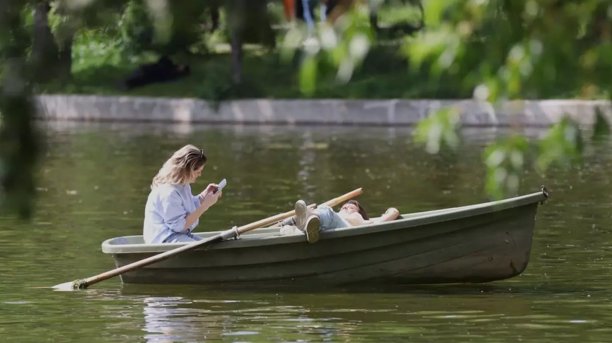 O tânără este într-o barcă pe apă și citește pe telefonul mobil, iar o alta stă culcată în barcă și se vede una dintre vâsle, în parcul Cișmigiu, București, 25 aprilie 2025