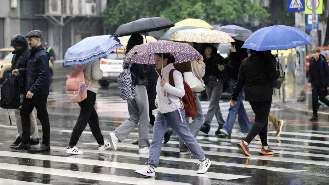 Mai mulți oameni cu umbrele în mâini și îmbrăcați cu geci traversează strada pe trecerea de pietoni, în București, într-o zi ploioasă și rece, 26 mai 2025