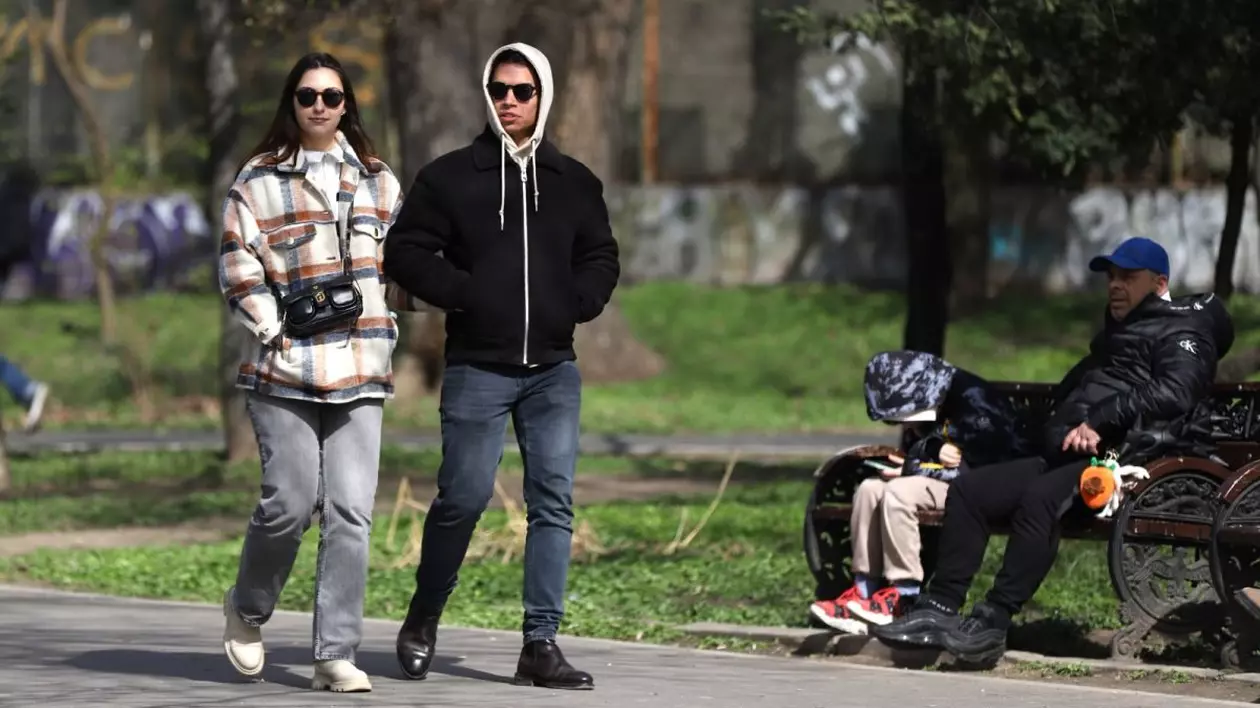 O femeie și un bărbat, îmbrăcați cu hanorace și glugă, se plimbă pe o alee din parc, iar un bărbat cu un copil stau pe o bancă, în parcul Cișmigiu, București, 19 martie 2025