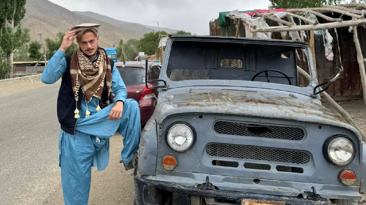În imagine un bărbat îmbrăcat în haine albastre stă cu piciorul rezemat de mașină pe o stradă din Afganistan