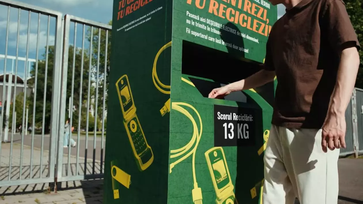 In imagine o persoane introduce un obiect electronic mic într-un container de reciclare verde, cu telefoane galbene desenate pe el, in spate se vede o poartă catre stadion