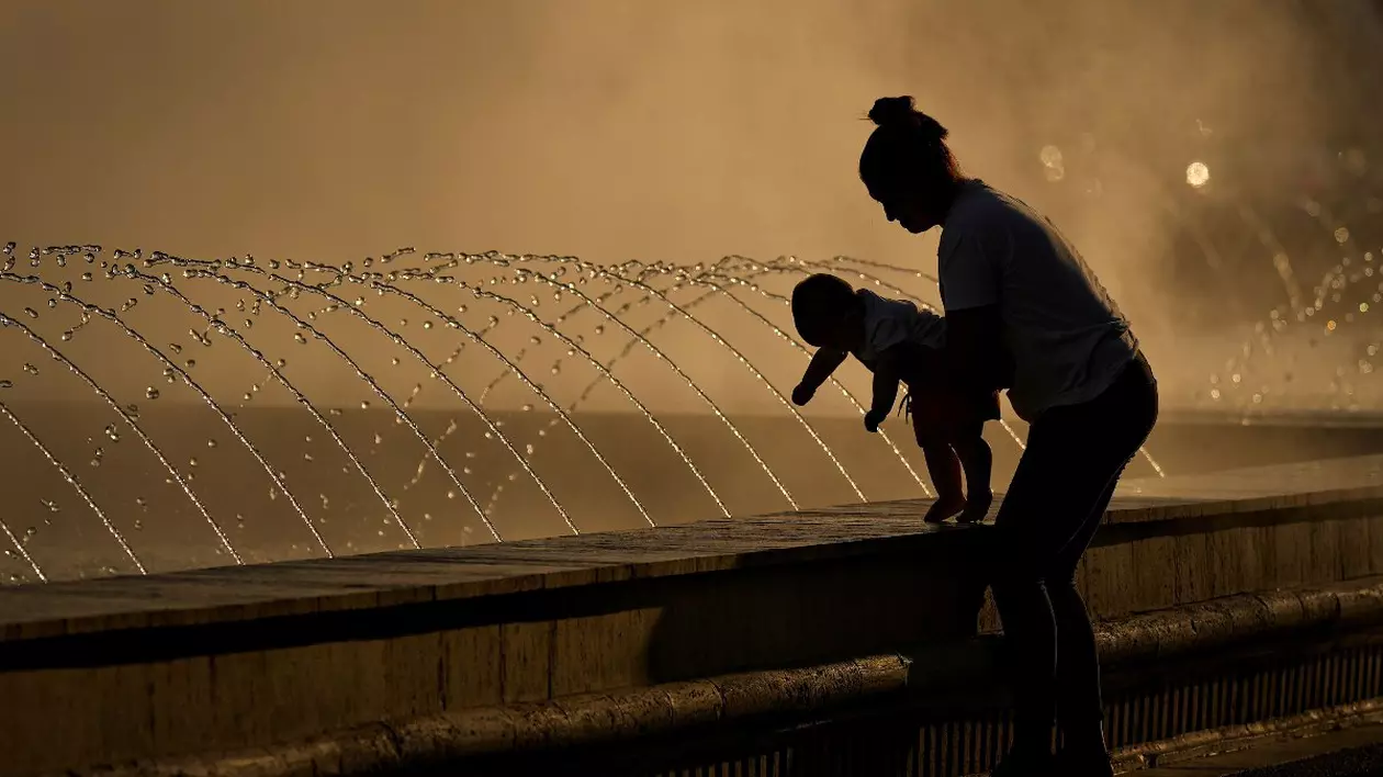 O femeie tine in brate un copil langa o perdea de vapori de apa, intr-o zi caniculara, la fantanilie de la Piata Unirii din Bucuresti, miercuri, 20 iulie 2022.