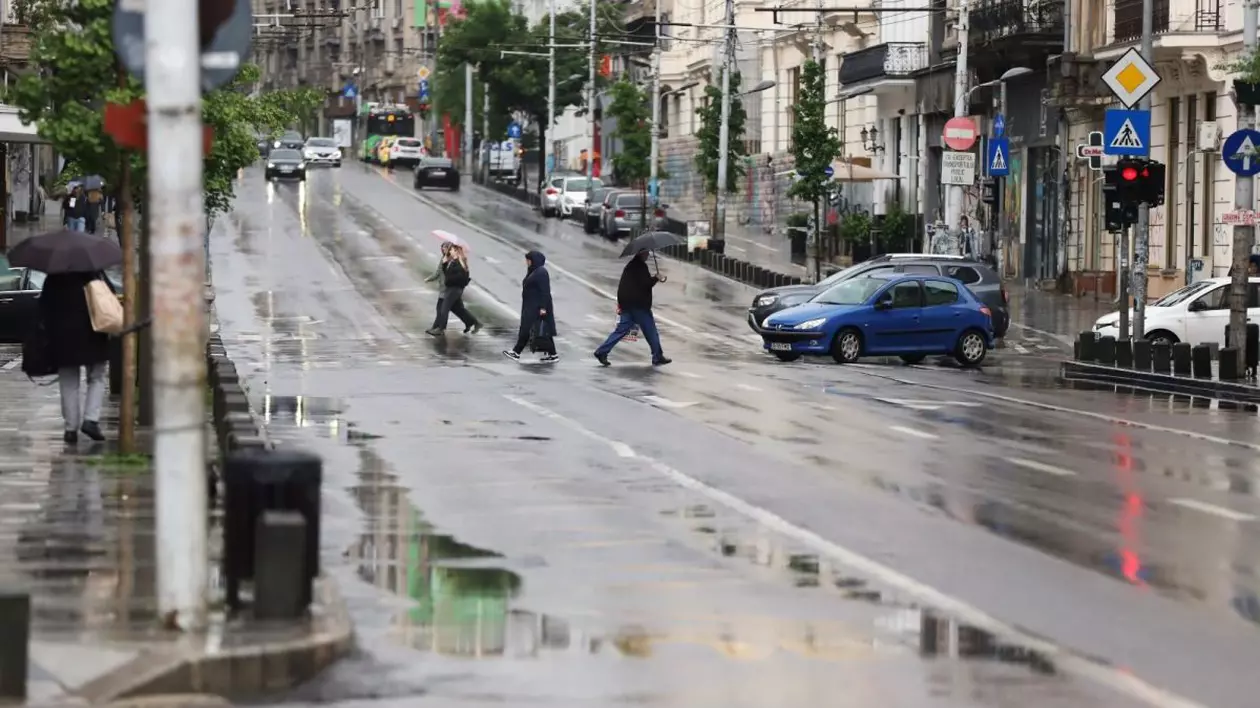 Mai mulți oameni cu umbrele în mâini traversează strada pe la trecerea de pietoni, câteva mașini sunt oprite, iar pe trotuar sunt alți oameni cu umbrele, într-o zi ploioasă de vară, zona Cișmigiu, București, 26 mai 2025