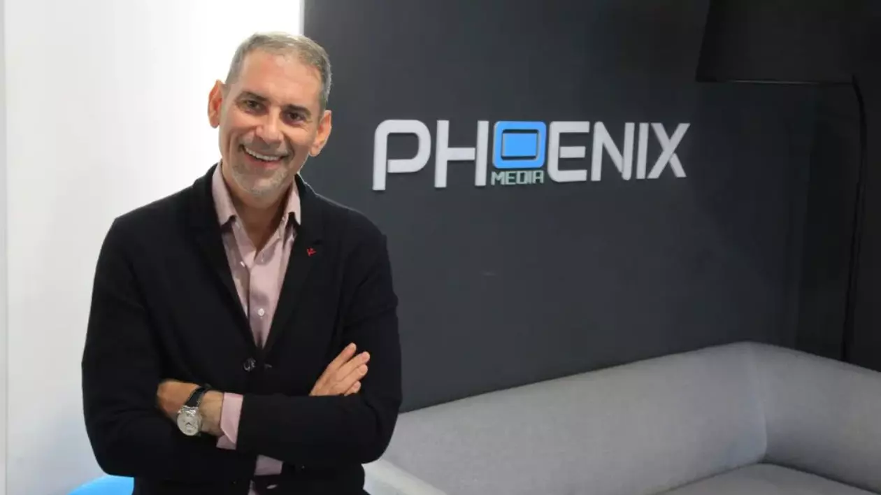 Dio Boacă, îmbrăcat cu un sacou negru și cămașă, iar pe fundal un perete cu logo-ul Phoenix Media