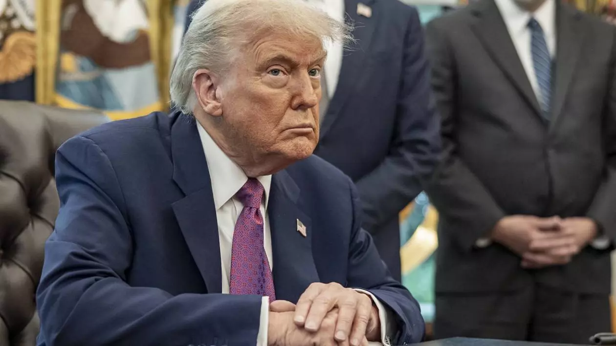 Președintele american Donald Trump în timp ce stă la biroul său de la Casa Albă, are mâinile încrucișate pe birou, poartă un costum albastru și o cravată mov.
