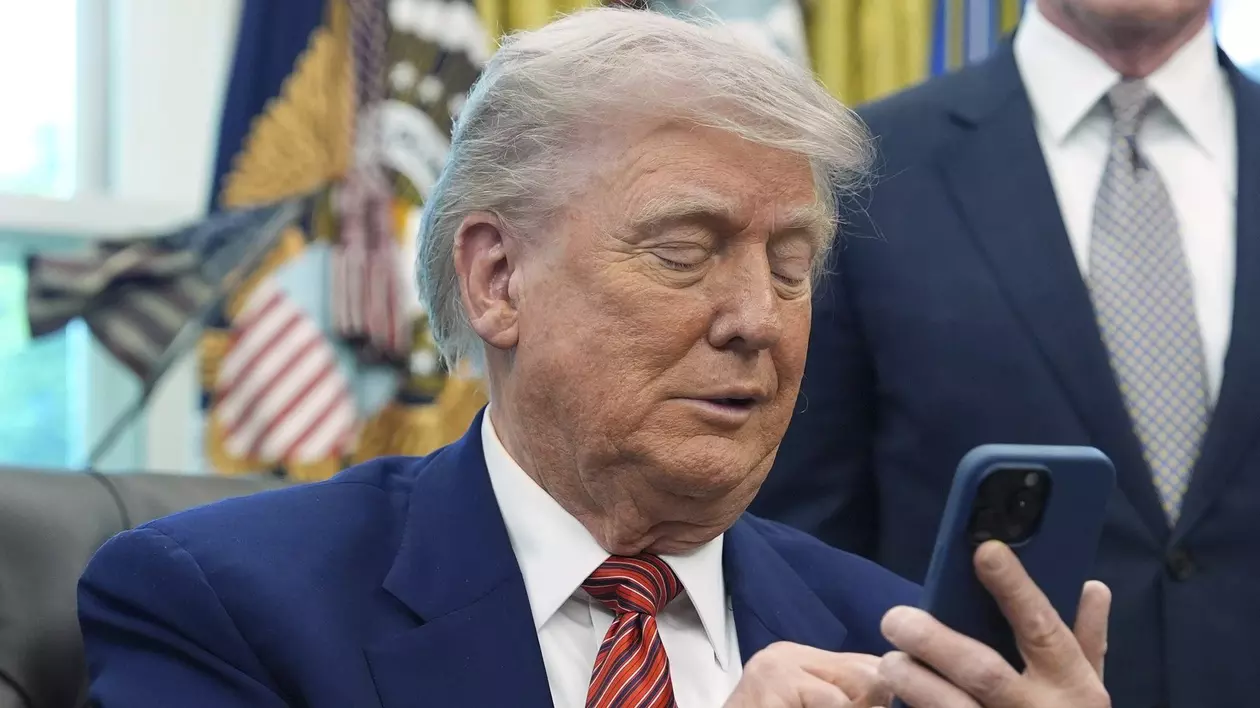 Donald Trump surprins în Biroul Oval în timp ce butonează telefonul, în mai 2025. Președintele SUA poartă un sacou albastru, o cămașă albă și o cravată roșie cu dungi negre. Acesta ține într-o mână telefonul, în timp ce pe cealaltă o folosește pe ecranul telefonului. Smartphone-ul are o husă albastră